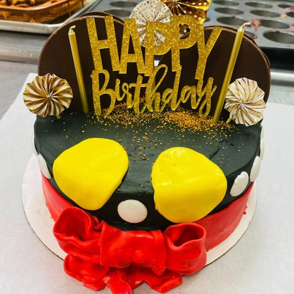 Mickey Mouse themed birthday cake with black frosting, yellow ears, red bow, and "Happy Birthday" topper.