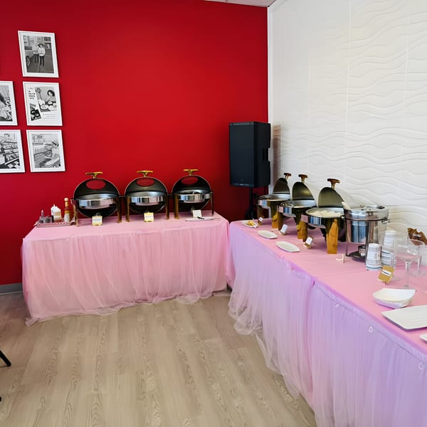 Filipino buffet setup with chafing dishes on pink-skirted tables against a red and white wall.