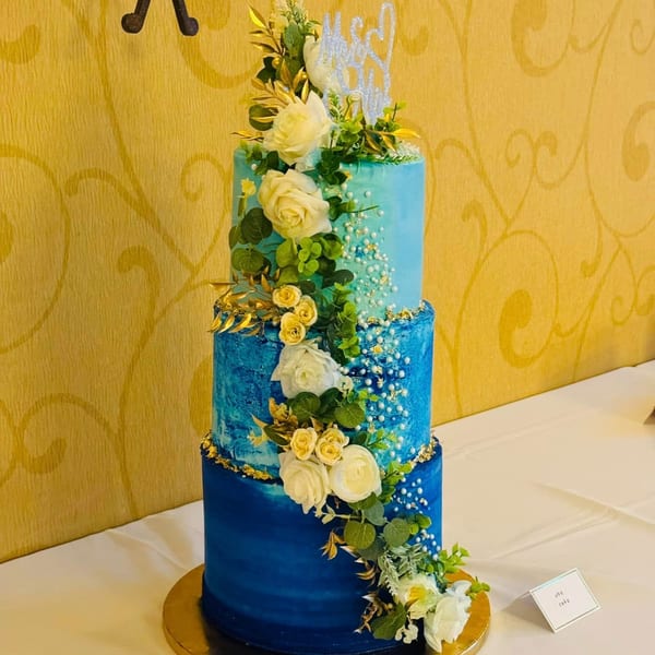 A three-tiered blue ombre Ube cake with white and yellow flowers, green leaves, and gold accents.