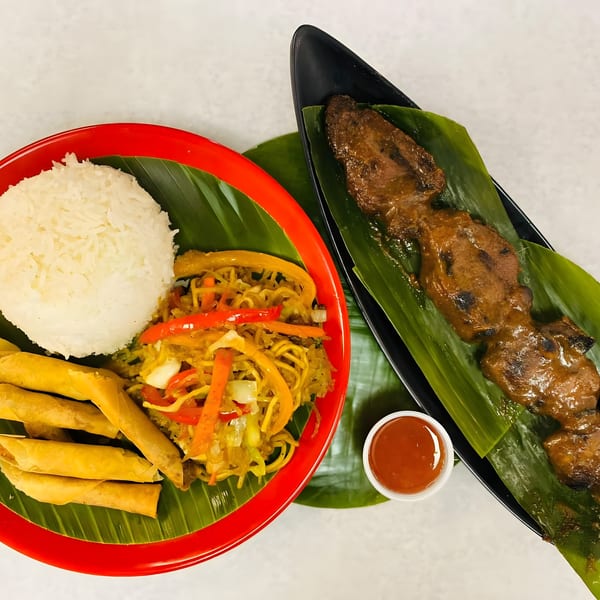 Filipino meal with rice, pancit, lumpia, and barbecue skewers on banana leaves.