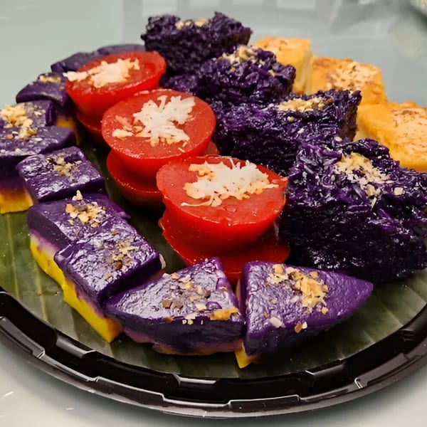Filipino desserts on a banana leaf: purple ube cakes, red puto, and yellow squares, all topped with nuts.